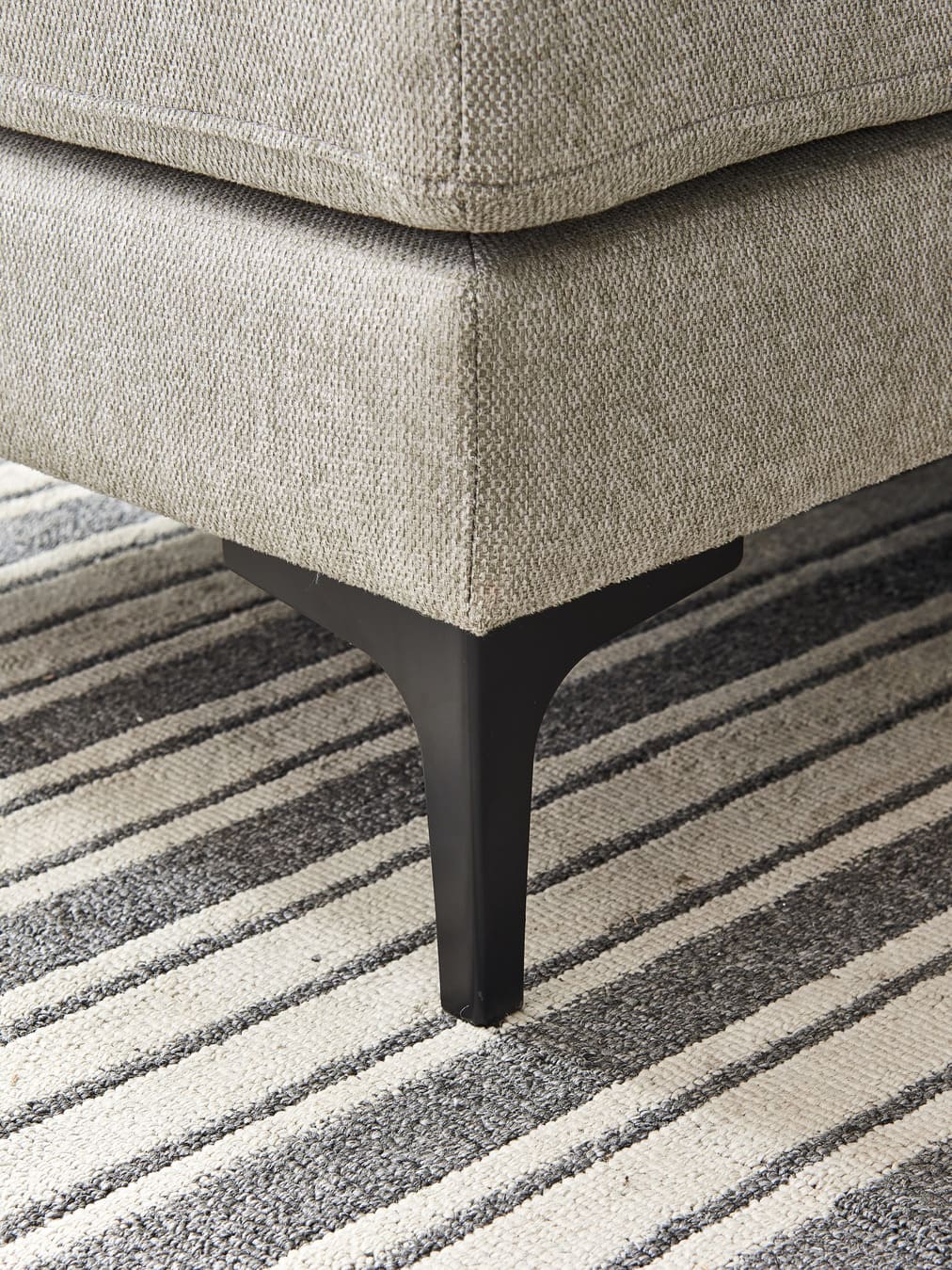 Close-up detail of Anek grey fabric sofa leg and corner on striped rug, showing textured upholstery and black metal feet